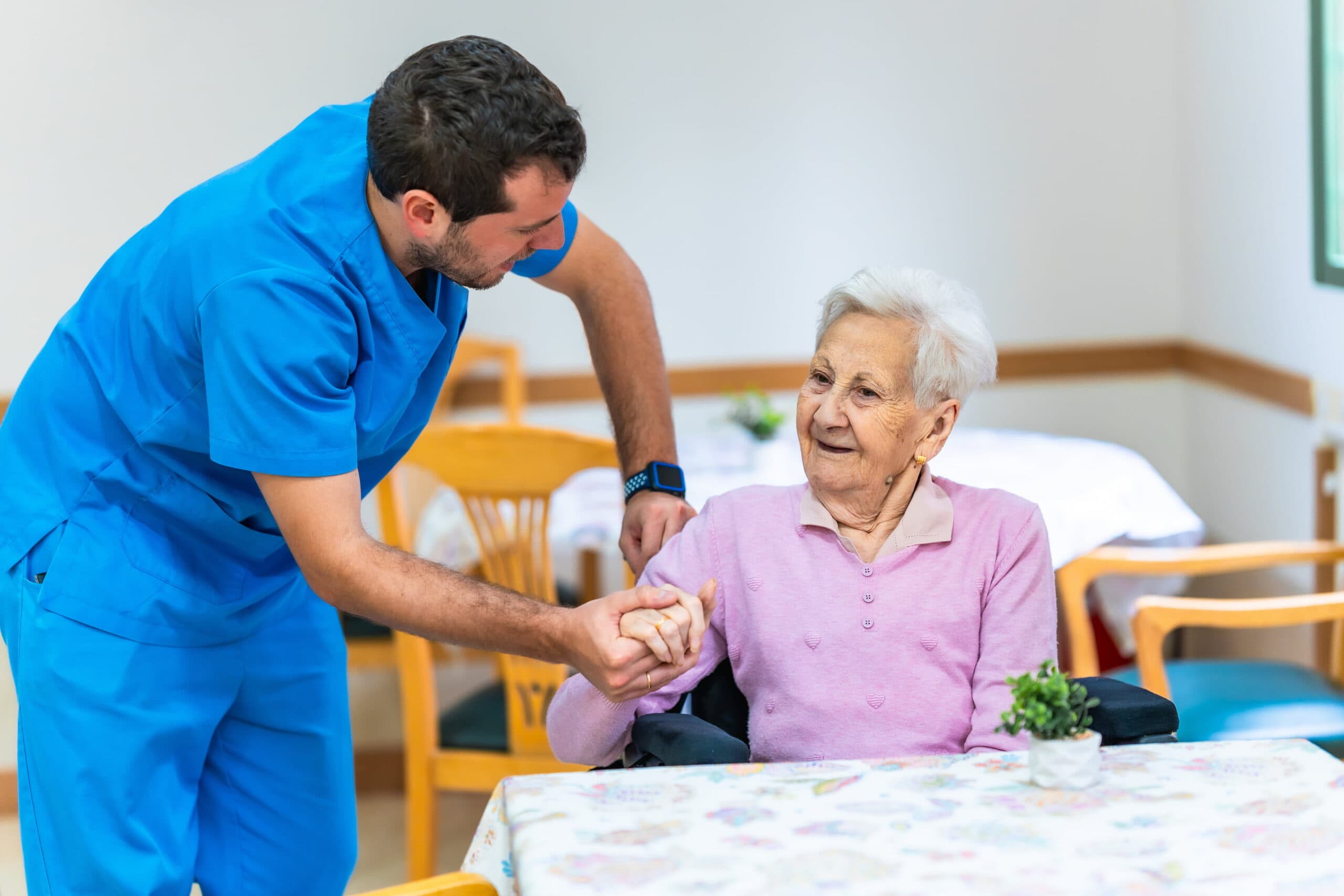 Male nurse in blue scrubs holding hands with elderly woman in a long term care facility