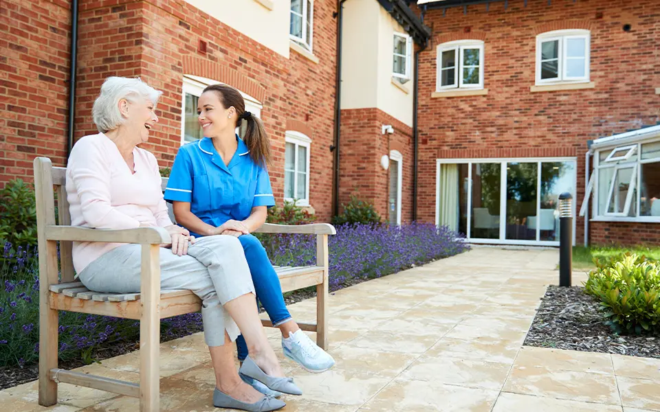 Senior Woman Sitting On Bench And Talking With Nurse In an Assisted Living Facility
