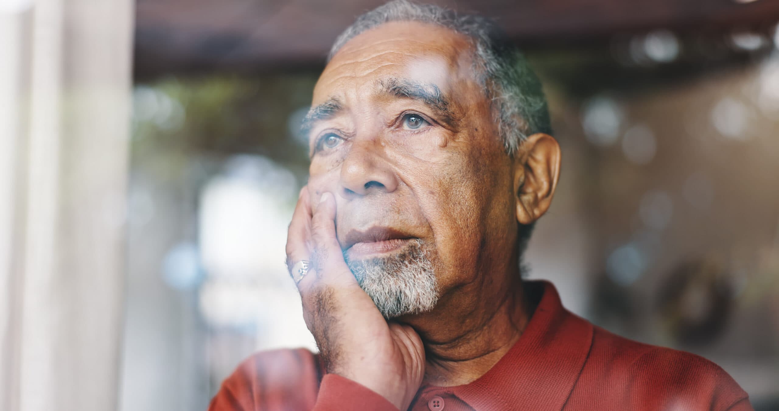 A senior man looking out the window with a look of distress while the sun is setting