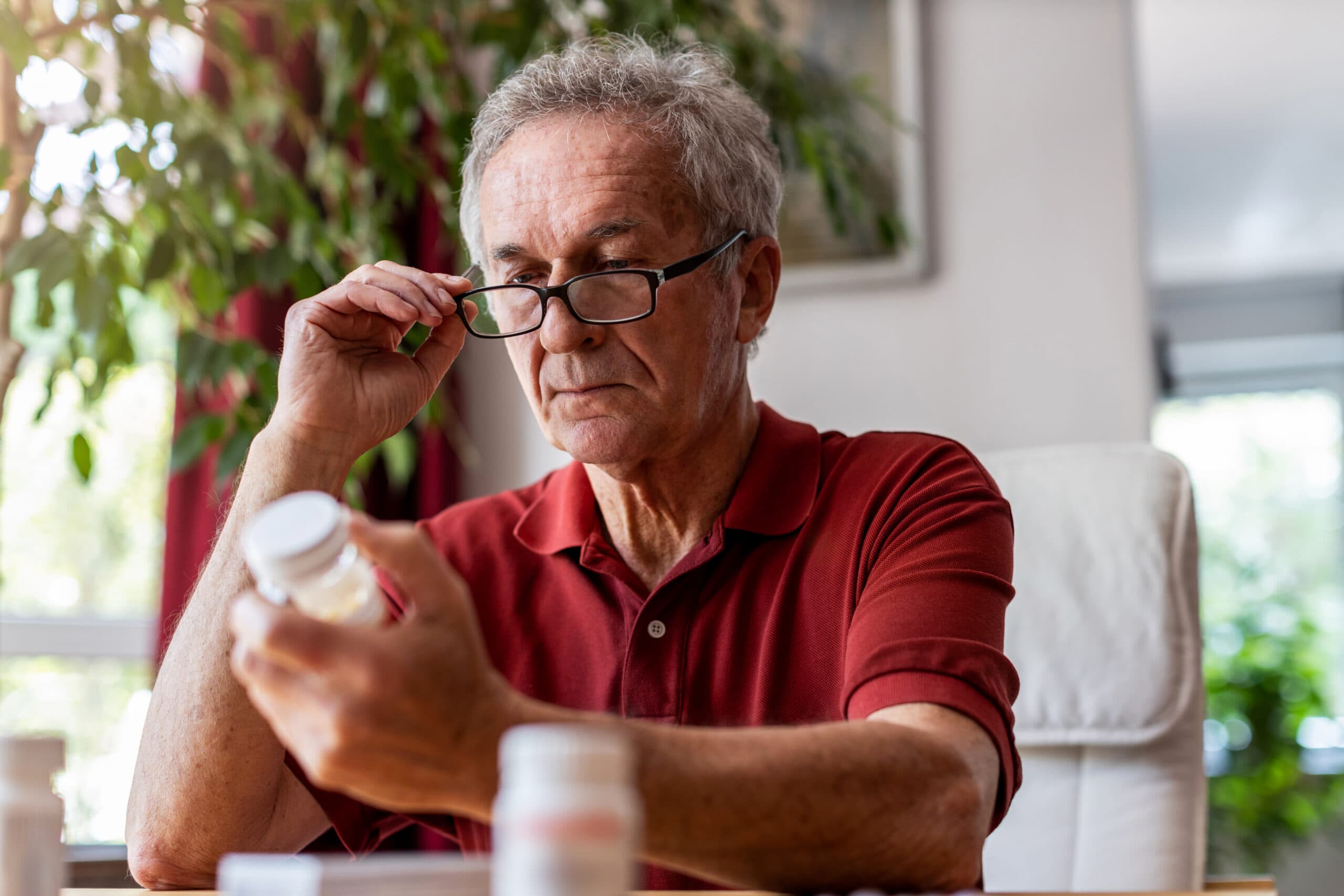 Image of an older man wearing a red shirt and black glasses reading the side of a prescription bottle